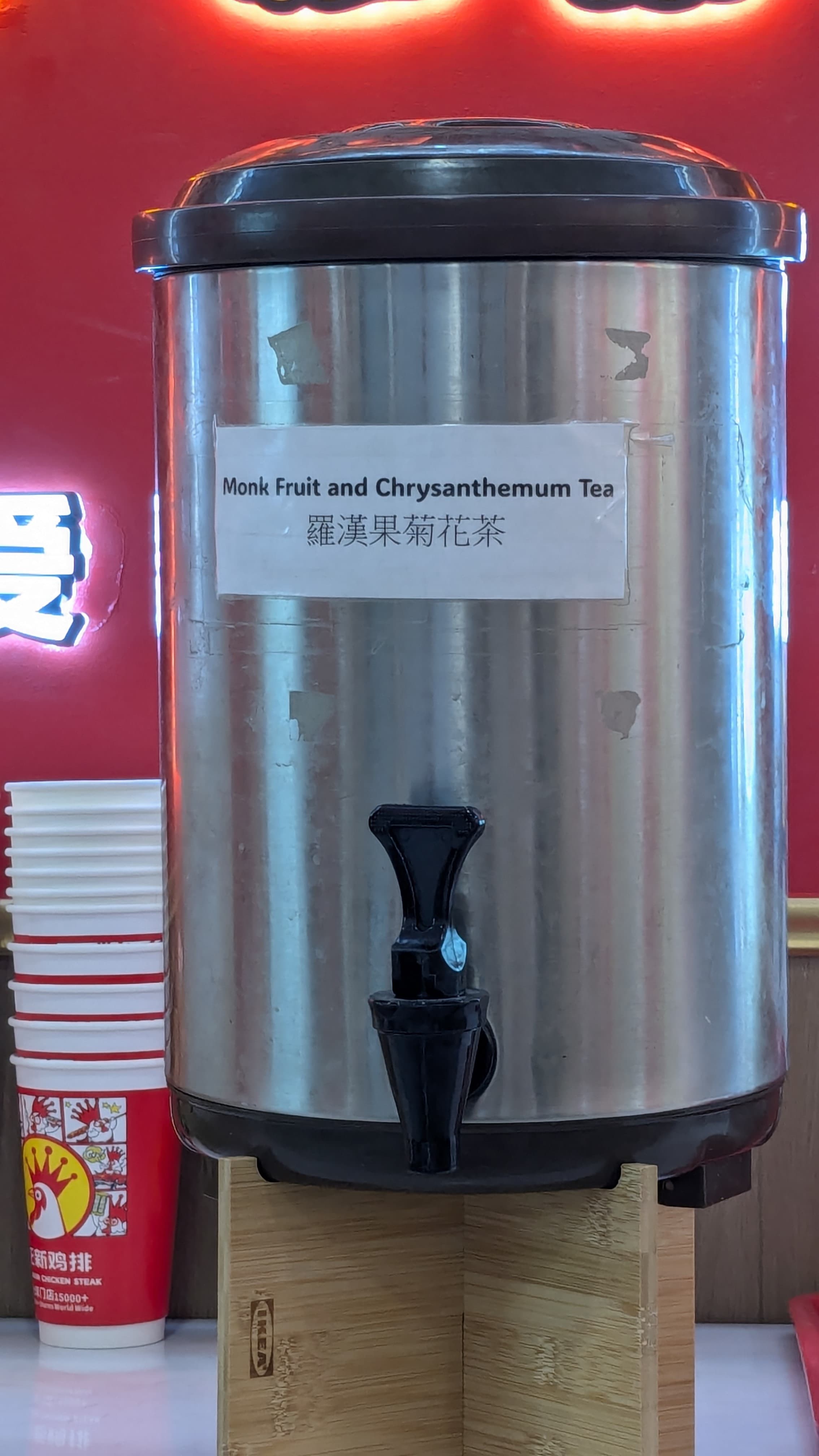 Stainless steel beverage dispenser labeled ‘Monk Fruit and Chrysanthemum Tea,’ with a black spout and stacked paper cups beside it.
