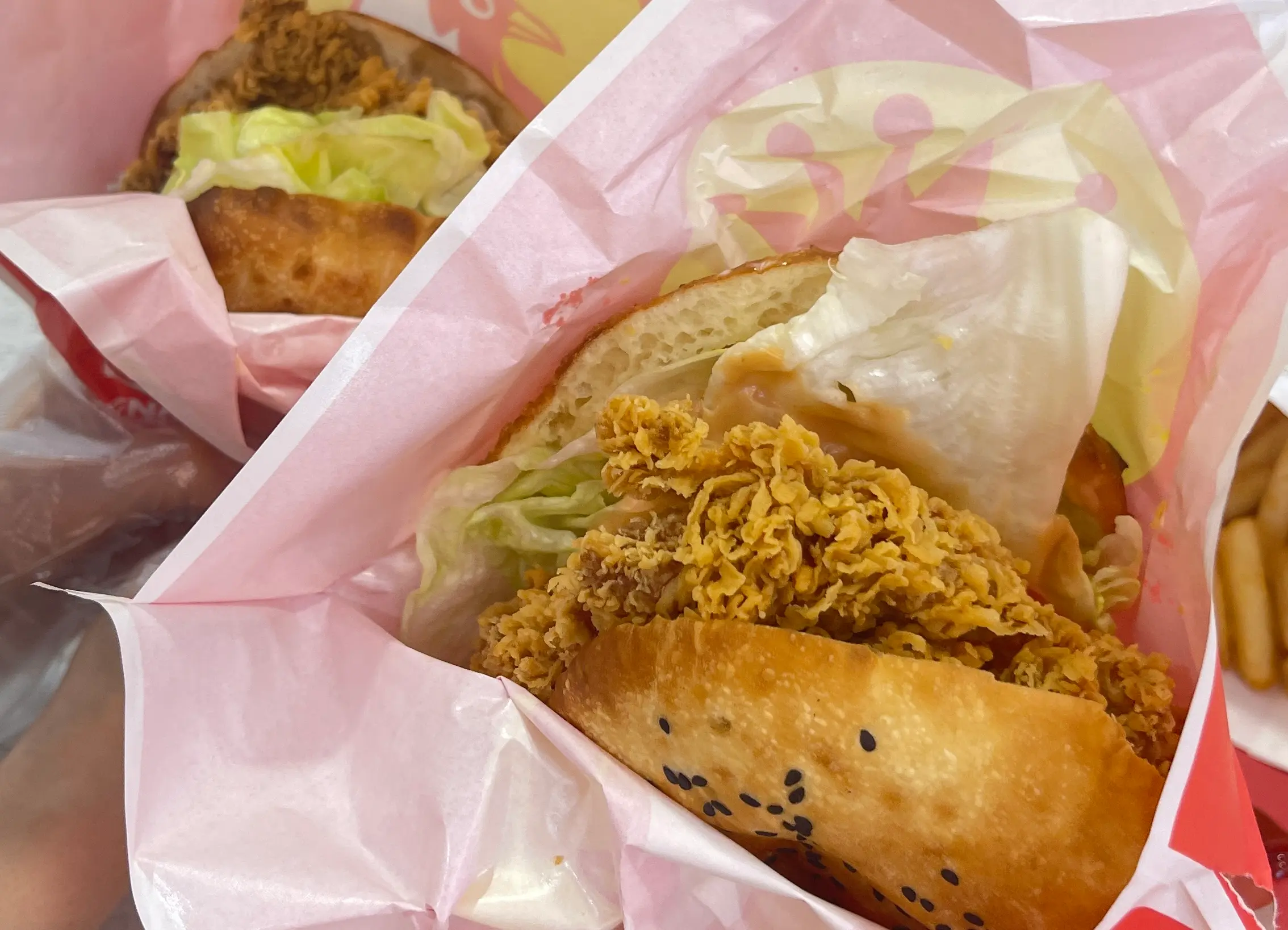 Close-up of a crispy fried chicken sandwich with lettuce, wrapped in pink paper, showing a golden bun and crunchy chicken filling.