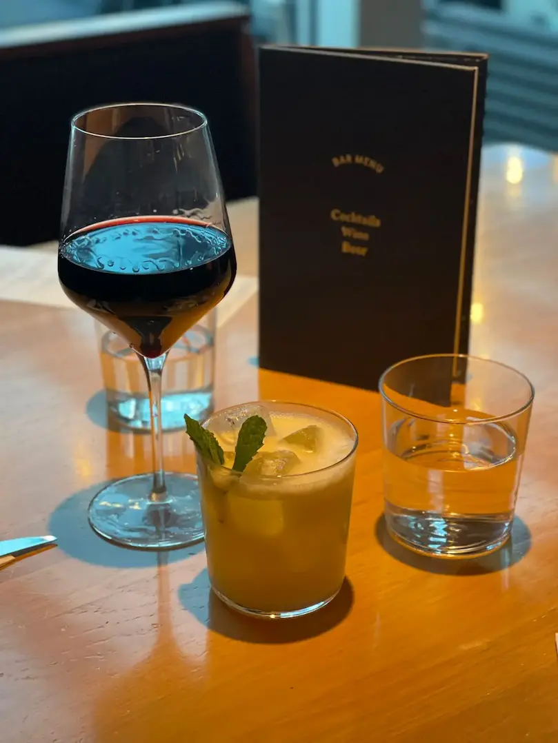 A glass of red wine and a yellow cocktail with mint on a wooden table, placed beside a menu and water glasses in a softly lit restaurant setting.