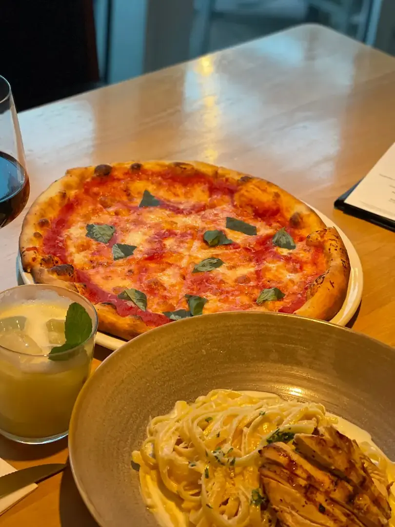 Margherita-style pizza topped with basil and a creamy pasta dish with grilled chicken, served on a wooden table with drinks in a restaurant.