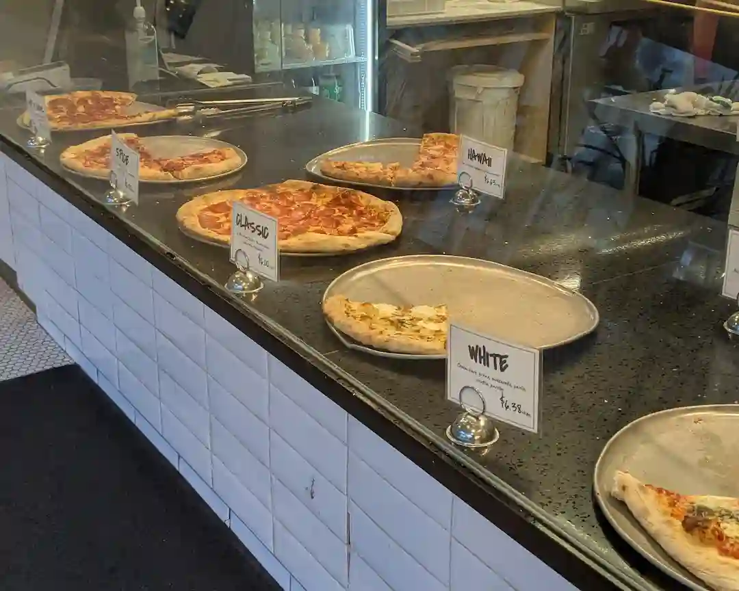 Pizza slices displayed behind a glass counter at Brooklyn Pizza, featuring classic pepperoni, cheese, and white slices on metal trays.