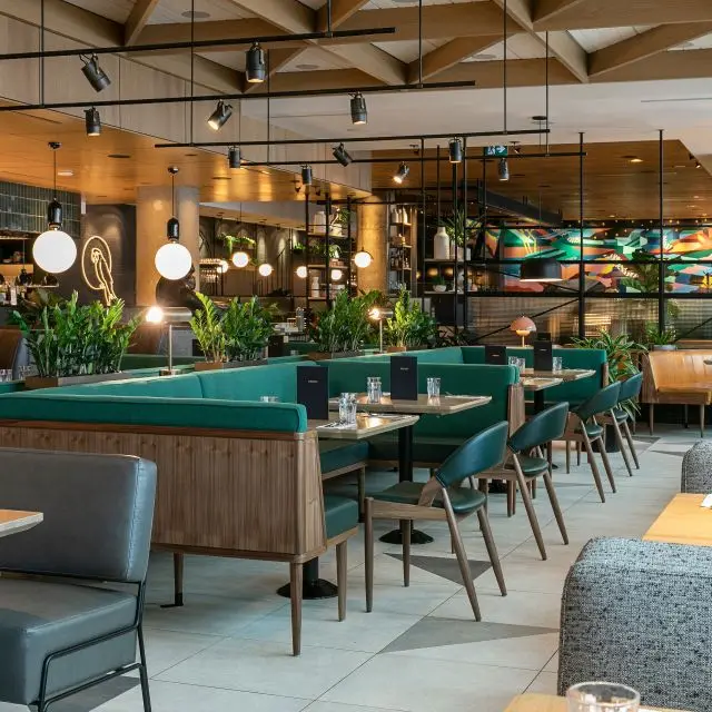 Modern restaurant interior with green booth seating, wooden tables, contemporary lighting, and indoor plants creating a stylish dining space.
