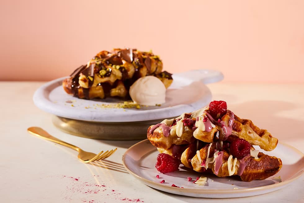 Plates of Belgian waffles topped with chocolate sauce, pistachios, raspberries, and cream, styled on a soft pastel background.