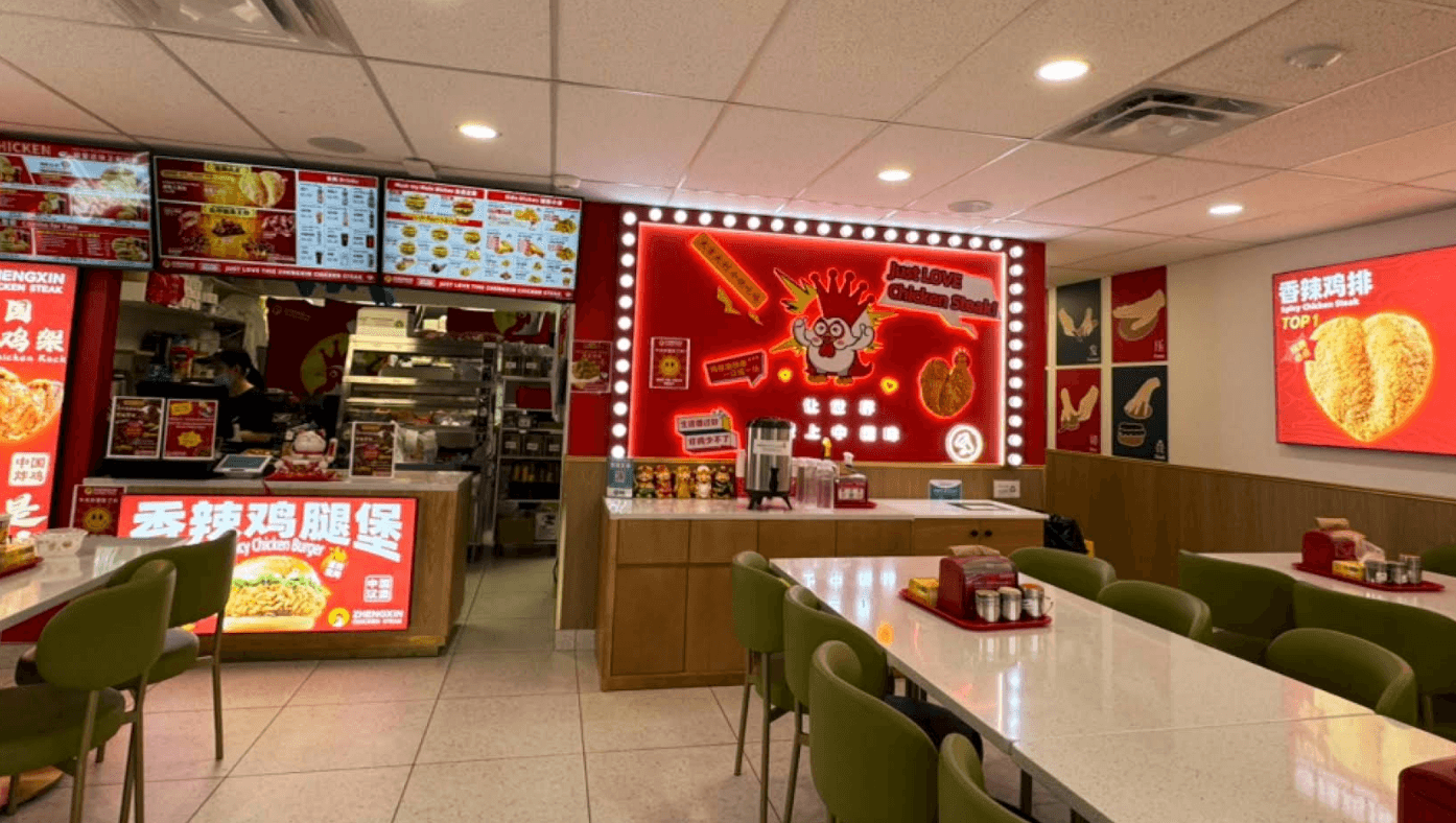 Bright red storefront of Zhengxin Chicken Steak featuring a large chicken statue outside, promotional posters on the windows, and an illuminated ‘OPEN’ sign.